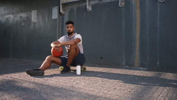 A sad young man with basketball ball outdoors in city, sitting and resting.