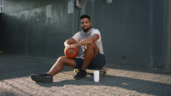 A happy young man with basketball ball outdoors in city, sitting and resting.