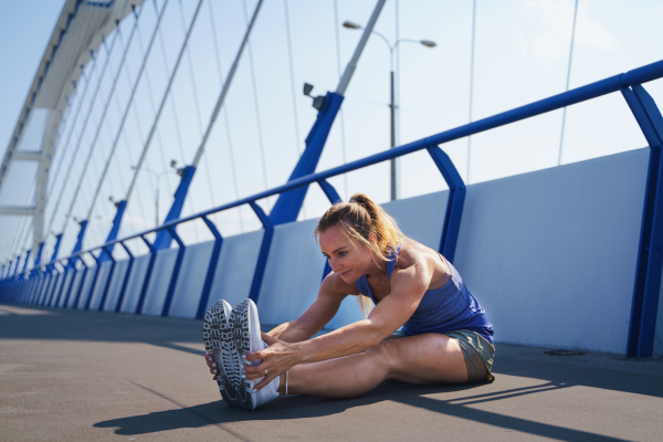 A mid adult woman runner stretching outdoors on bridge in city, healthy lifestyle concept.