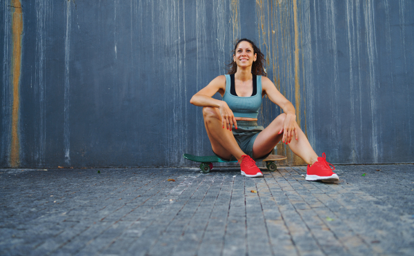 A front view portrait of mid adult woman sitting on skateboard outdoors in city, resting.