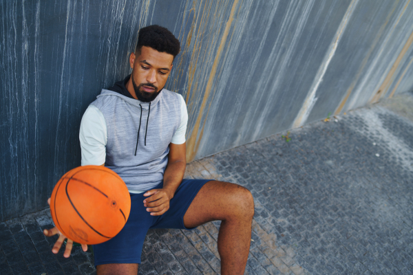 A young man with basketball doing exercise outdoors in city, sitting and resting.