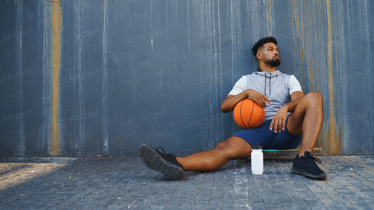 A young man with basketball doing exercise outdoors in city, sitting and resting.