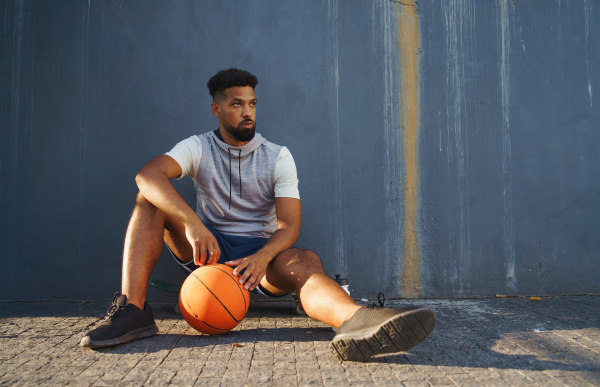 A young man with basketball doing exercise outdoors in city, sitting and resting.