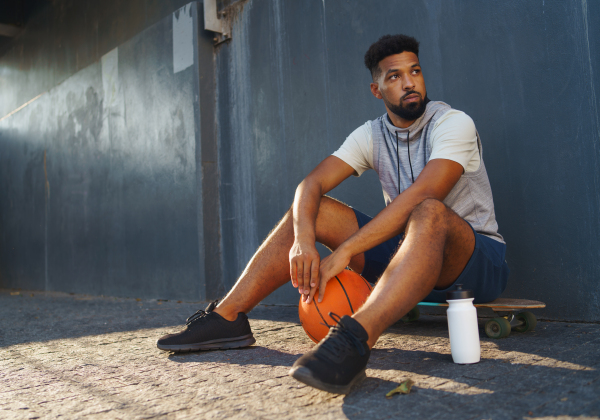 A sad young man with basketball ball outdoors in city, sitting and resting.