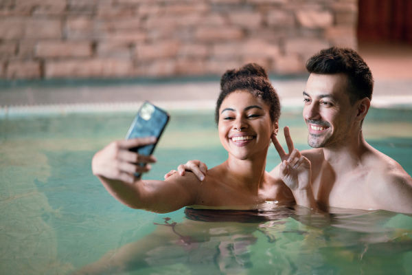 Portrait of happy young couple in indoor hot spring thermal pool, taking selfie.