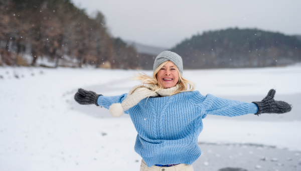 A front view portrait of cheerful senior woman with hat and mittens outdoors having fun in snowy nature.