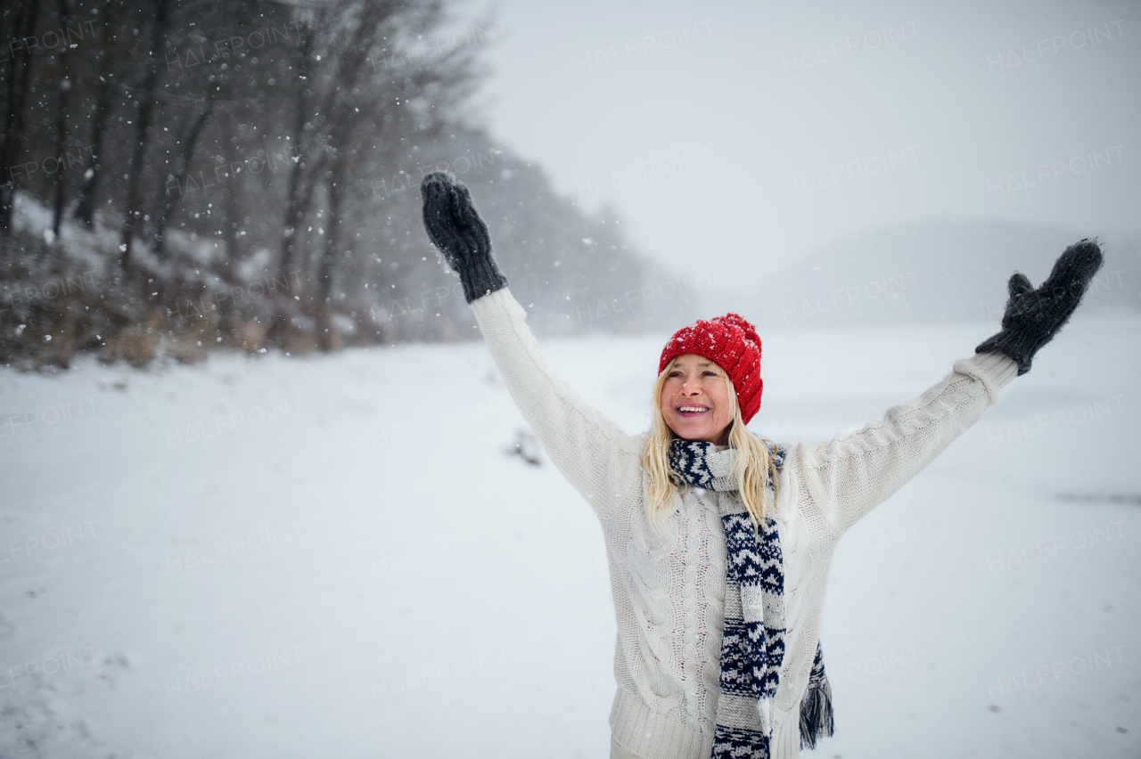 Front view portrait of happy senior woman with hat and mittens outdoors standing in snowy nature.