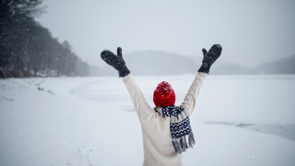 Rear view of an active senior woman on walk outdoors in snowy nature, walking by frozen lake with hands up.