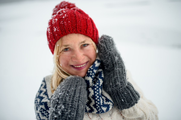 Headshot portrait of happy senior woman with hat and outdoors standing in snowy nature, looking at camera.