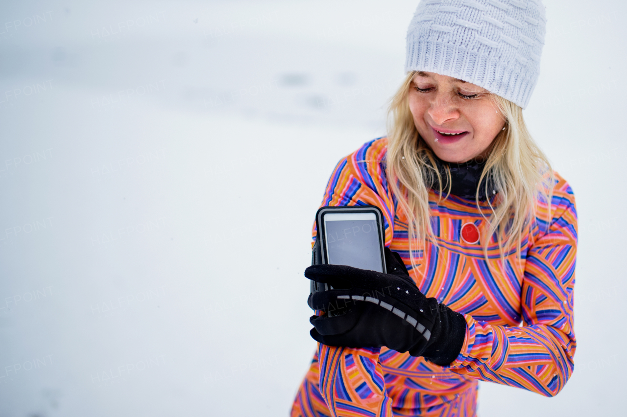 An active senior woman runner adjusting smartphone, attached to arm, for exercising outdoors in snowy winte.