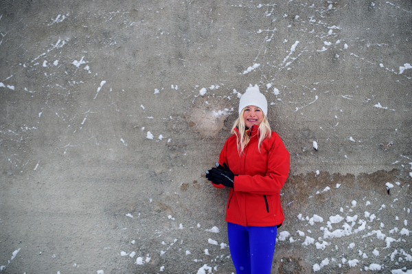 An active senior woman runner standing against concrete wall outdoors in snowy winter, looking at camera.