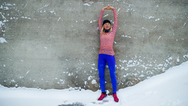 An active senior woman runner standing against concrete wall outdoors in snowy winter, stretching.