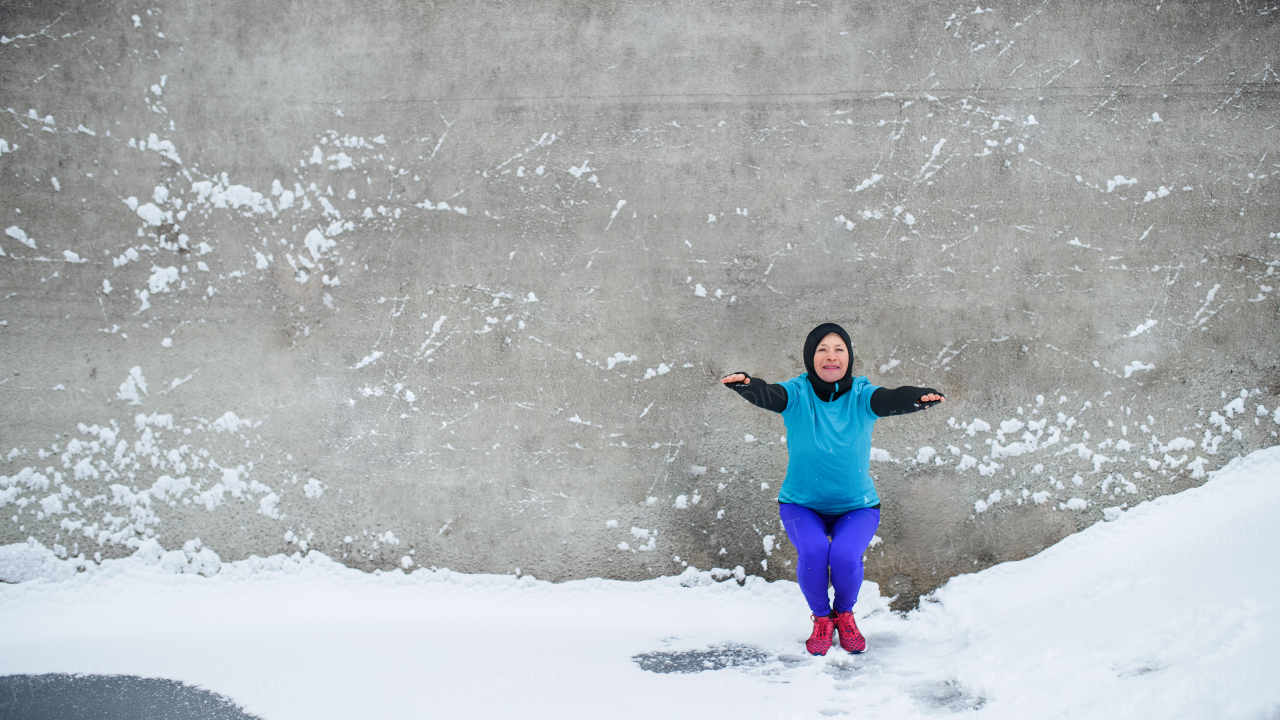 A front view of active senior woman outdoors in snowy winter, doing squats. Copy space.