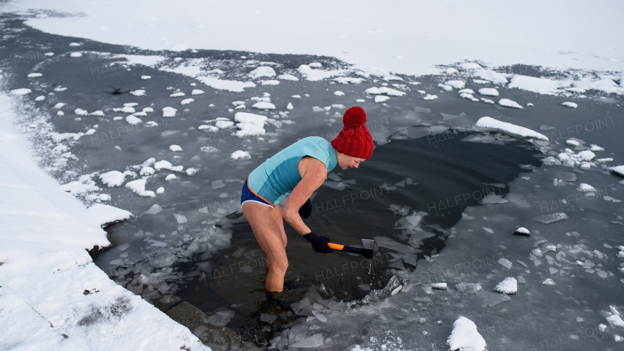 Happy active senior woman in swimsuit breaking ice with axe outdoors in winter, cold therapy concept.