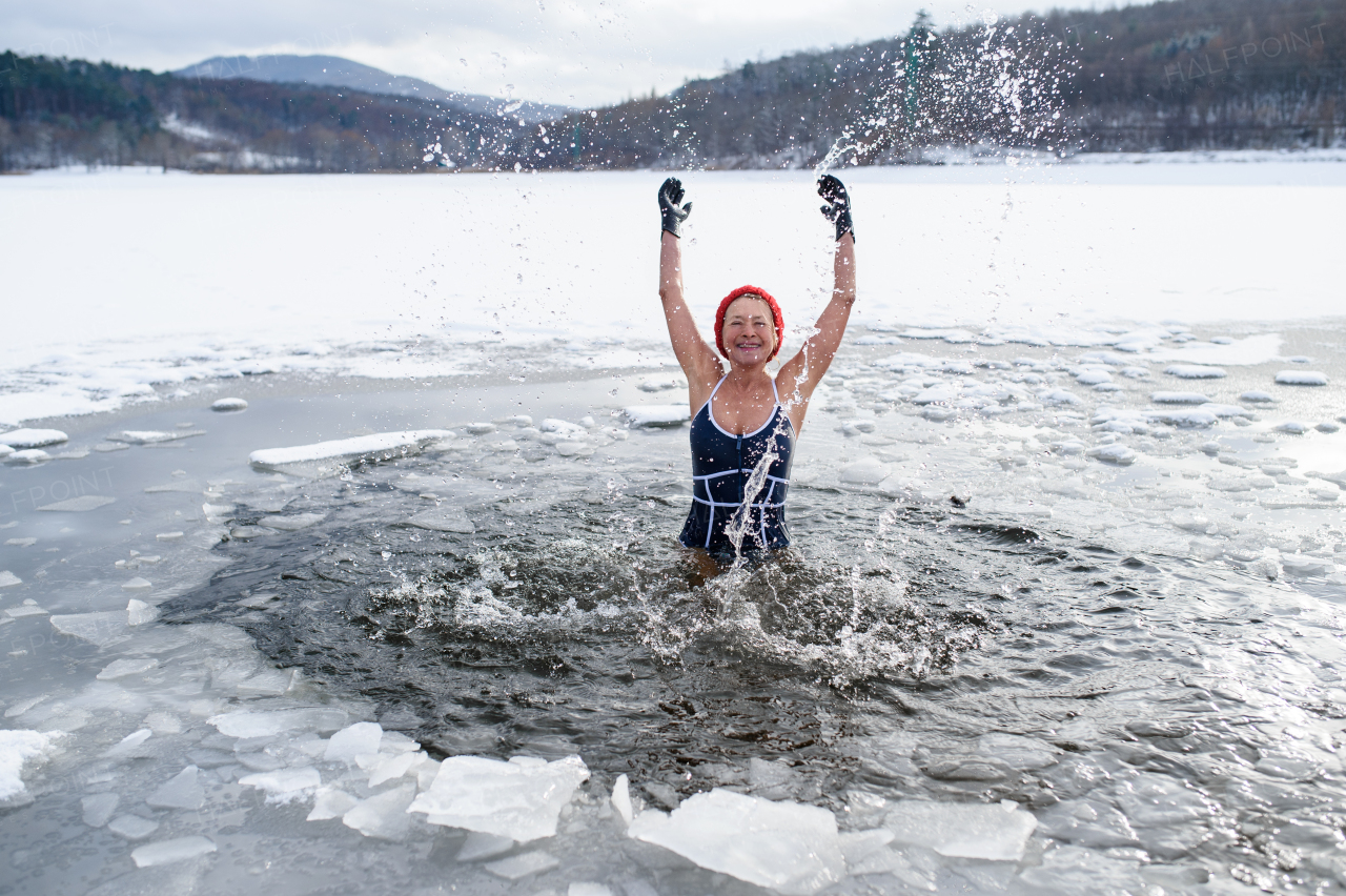 A front view of active senior woman in swimsuit splashing water outdoors in winter, cold therapy concept.