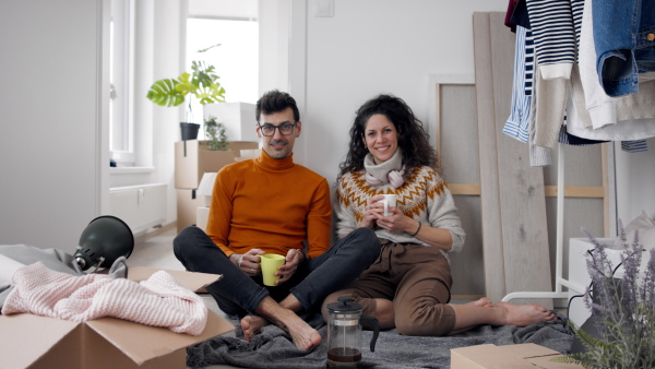 Happy young couple with boxes moving in new flat, having coffee, looking at camera. New home and relocation concept.