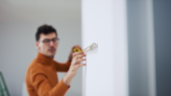 A young male owner in new flat, moving in, measuring with measuring tape. New home and relocation concept.