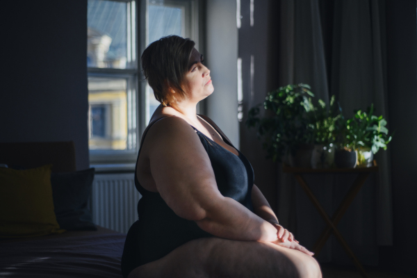 Depressed lonely fat woman sitting and looking through the window at home at dark.