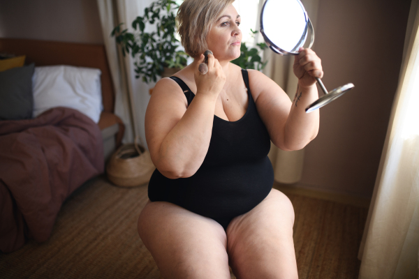 A fat woman sitting and looking at mirror at home, selfcare concept.