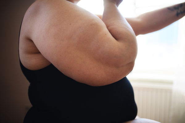 Close up for a womans obese body, standing and looking through the window at home.