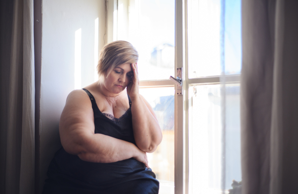 A depressed lonely fat woman in underwear sitting near window, looking down and thinking at home.