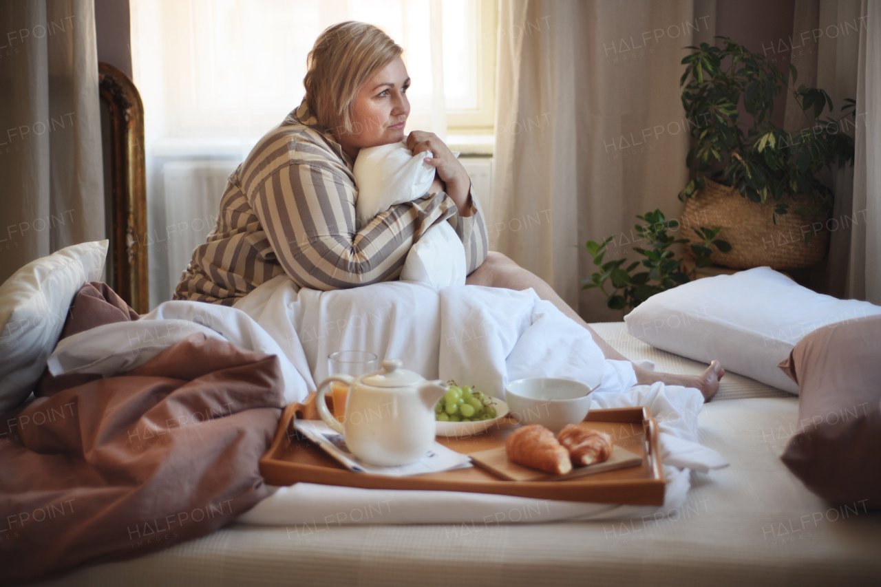 A happy overweight woman with headphones and smartphone having breakfast in bed at home.