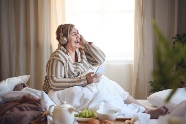 A happy overweight woman with headphones and smartphone having breakfast in bed at home.