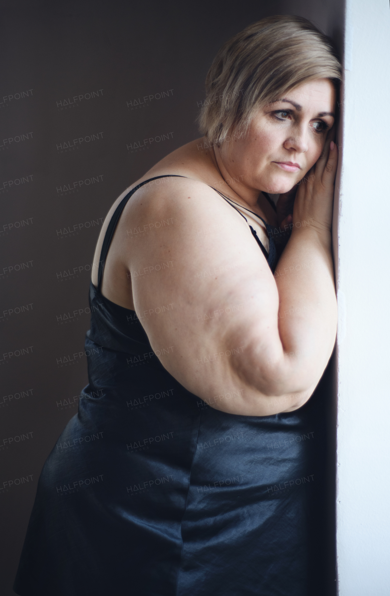 A depressed lonely fat woman standing and looking through the window at home.