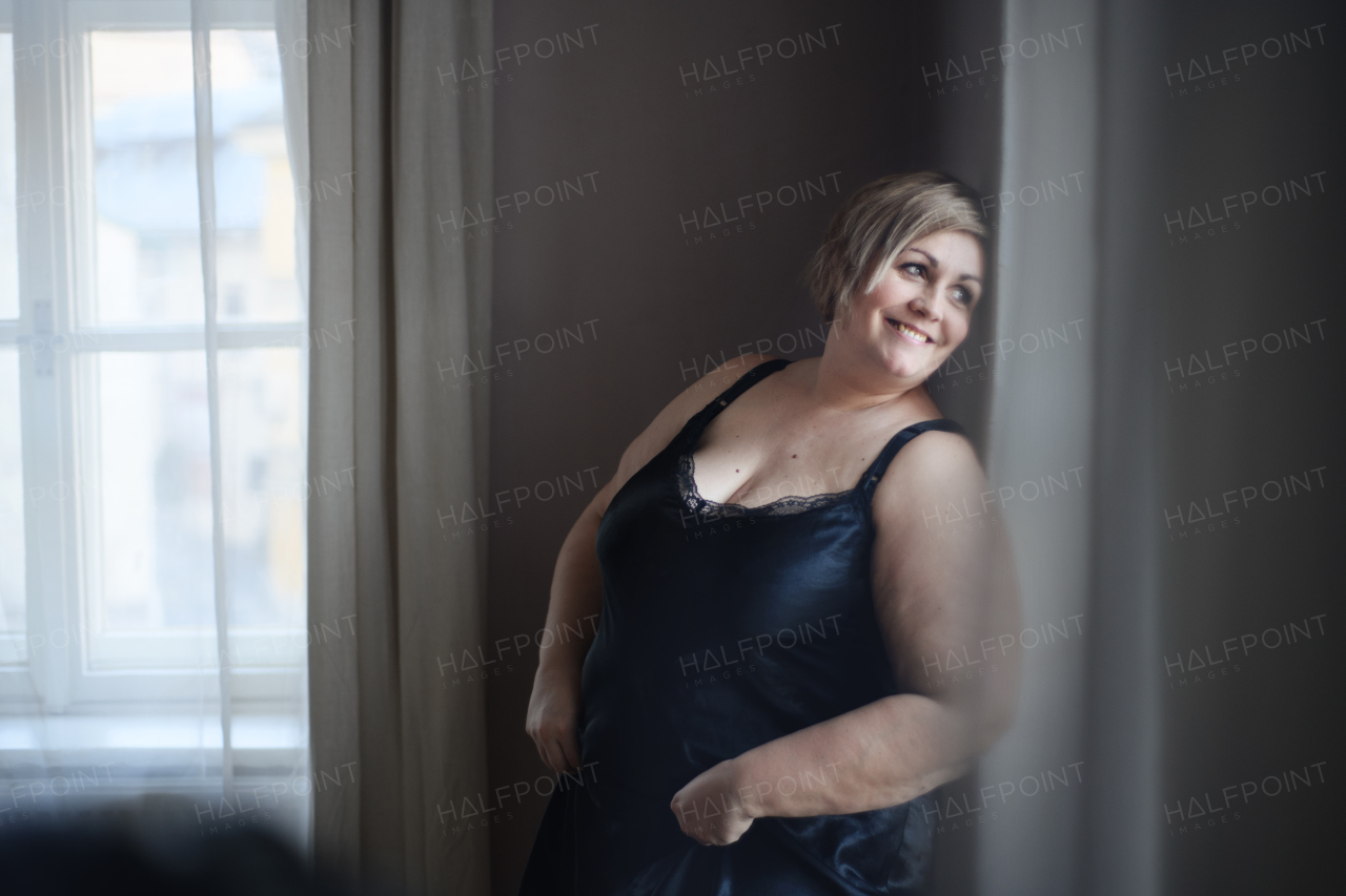 A happy overweight woman in underwear standing and looking out through window at home.