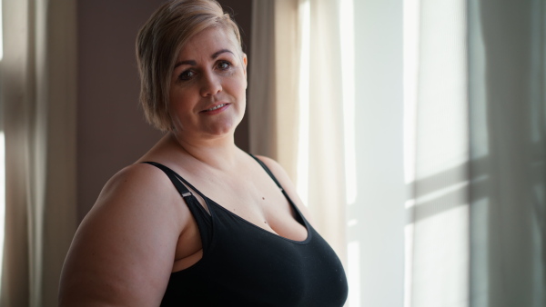 An overweight woman in underwear standing by window and looking at camera.