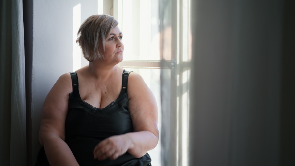 A depressed lonely fat woman sitting aby window at home and thinking.