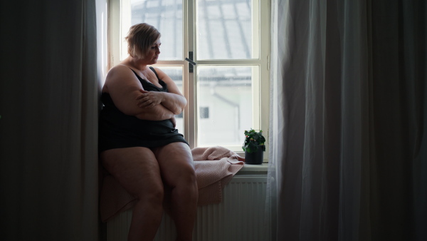 A depressed lonely fat woman sitting aby window at home and thinking.