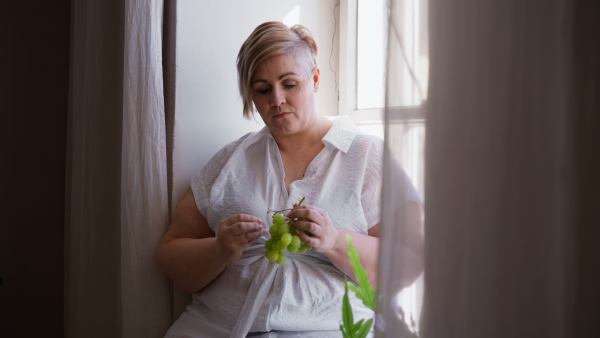 A depressed lonely fat woman sitting aby window, eating grapews and thinking at home.