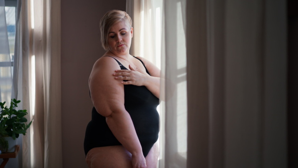 An overweight woman in underwear standing by window and looking at camera.