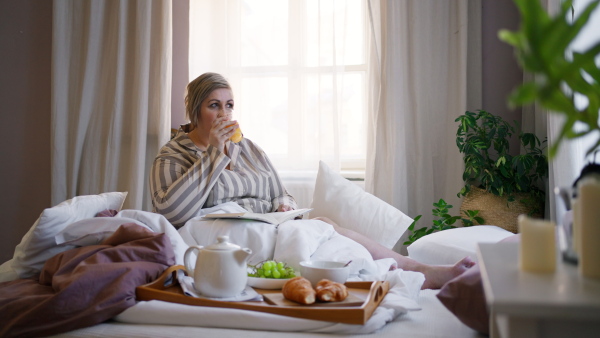 A happy overweight woman reading book and having breakfast in bed at home.