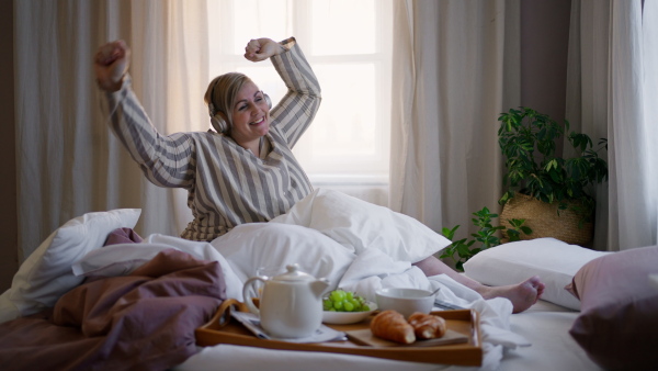 A happy overweight woman in pyjamas with headphones dancing when sitting in bed at home.