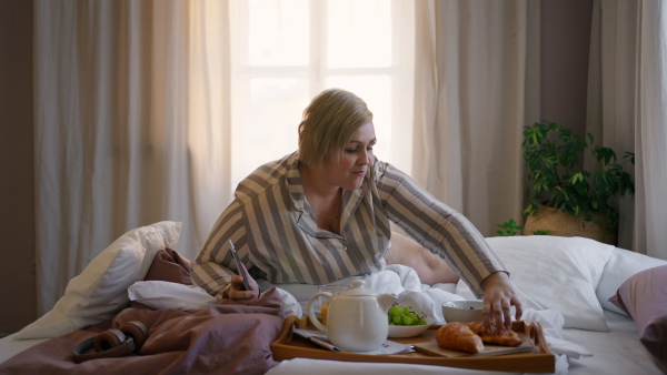 A happy overweight woman taking selfie and having breakfast in bed at home.