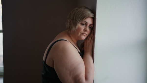 A depressed lonely fat woman standing and leaning the wall at home and thinking.