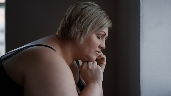 A depressed lonely fat woman standing and leaning the wall at home and thinking.