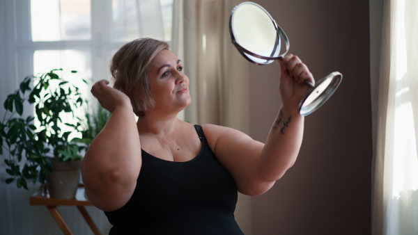 A fat woman sitting and looking at mirror at home, selfcare concept.