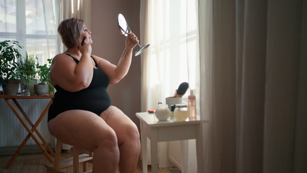 A fat woman sitting and looking at mirror and applying make up at home, selfcare concept.