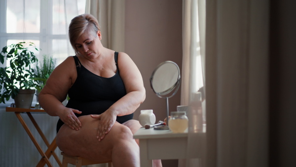 A fat woman sitting and applying cream on her legs at home, selfcare concept.