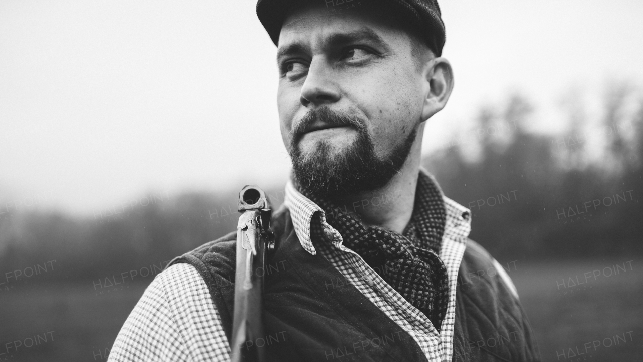 A portrait of hunter man in traditional shooting clothes on field holding shotgun, black and white photo.