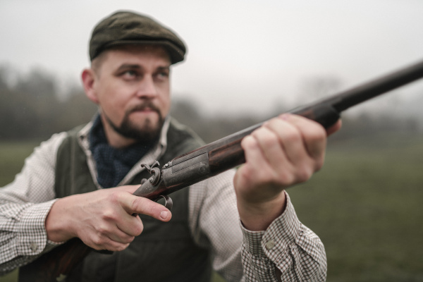 A hunter man in traditional shooting clothes on field aiming with shotgun.