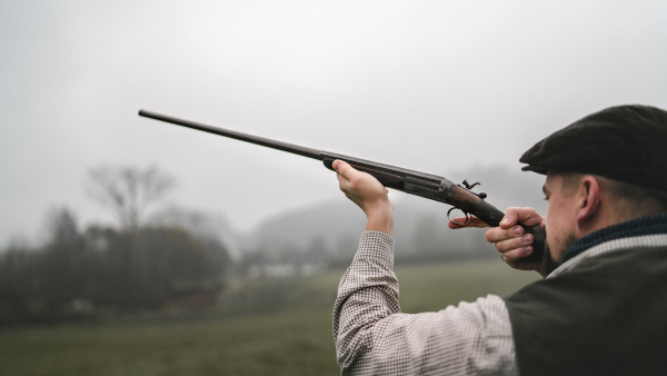 A hunter man in traditional shooting clothes on field aiming with shotgun.