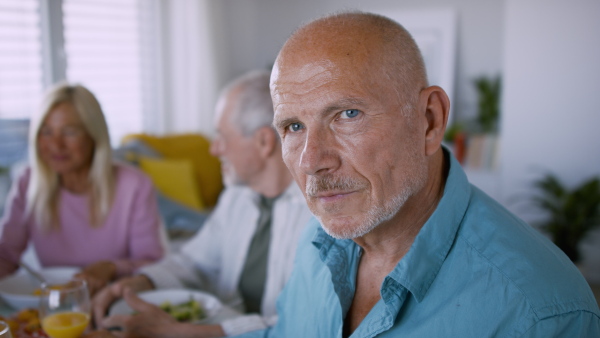 A senior man with friends having party indoors, eating at the table and looking at camera.