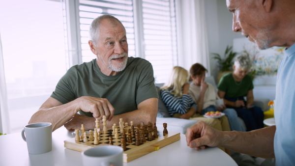 Two senior friends playing chess and talking indoors, social gathering concept.