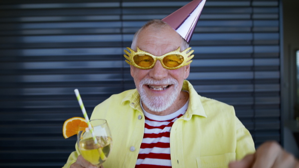 Crazy senior man with party hat and drink standing outdoors, taking selfie.