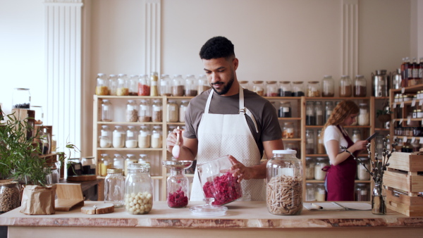 A young man filling jar by dryed raspberries in zero waste shop, looking at camera.