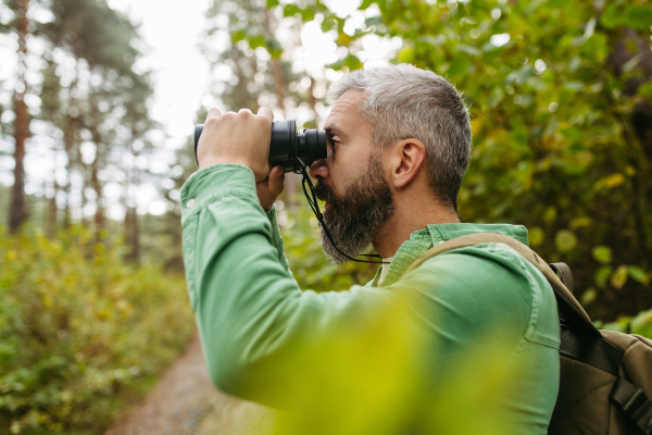 Handsome man watching wildlife with binoculars, enjoying peaceful atmosphere of forest, forestbathing.